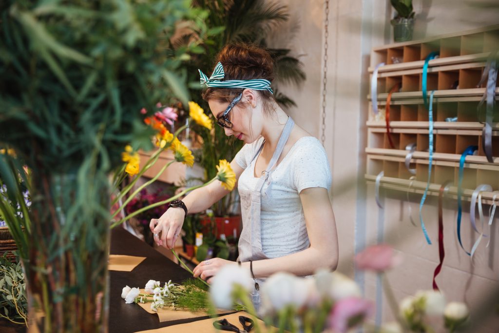 Leveraging Messaging to Inform and Engage - female florist trimming flowers for a bouquet, smiling in bright floral shop - overhead marketing concept showing that employees can focus on work while overhead messaging informs customers