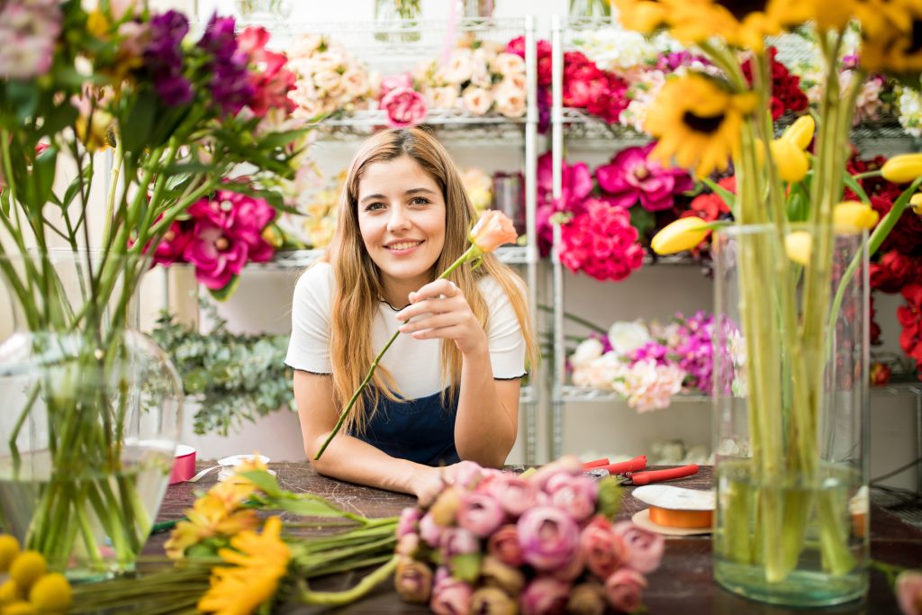 overhead marketing concept - florist in brightly lit shop holding flower and smiling at the camera - conveys idea that overhead messaging and music creates a positive environment for customers and employees