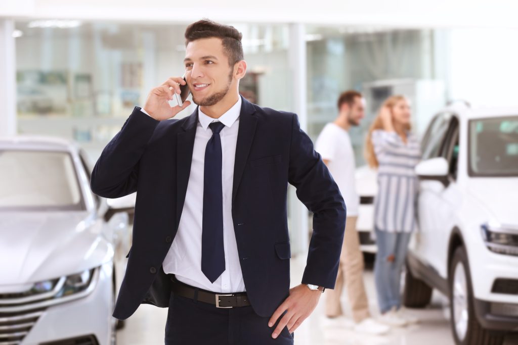 Leveraging On-Hold Marketing to Inform and Persuade - Car salesman standing in dealership showroom talking on phone, smiling, with couple looking at cars in the background - concept for the importance of on-hold marketing in a car dealership