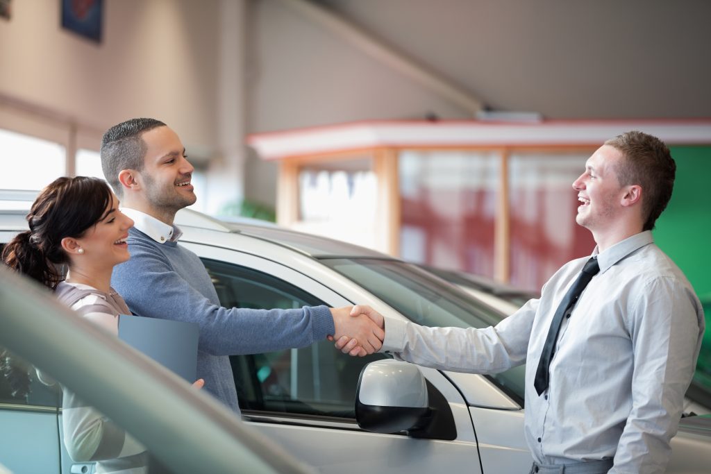 Best Practices for On-Hold Music and Messaging - smiling car salesman shaking hands with man in showroom while woman smiles holding folder of car paperwork - successful on-hold marketing concept 