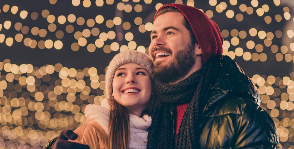 Happy smiling couple in winter clothes looking around, to convey the ideas that they are at a winter holiday event and listening to holiday music, surrounded by Christmas lights