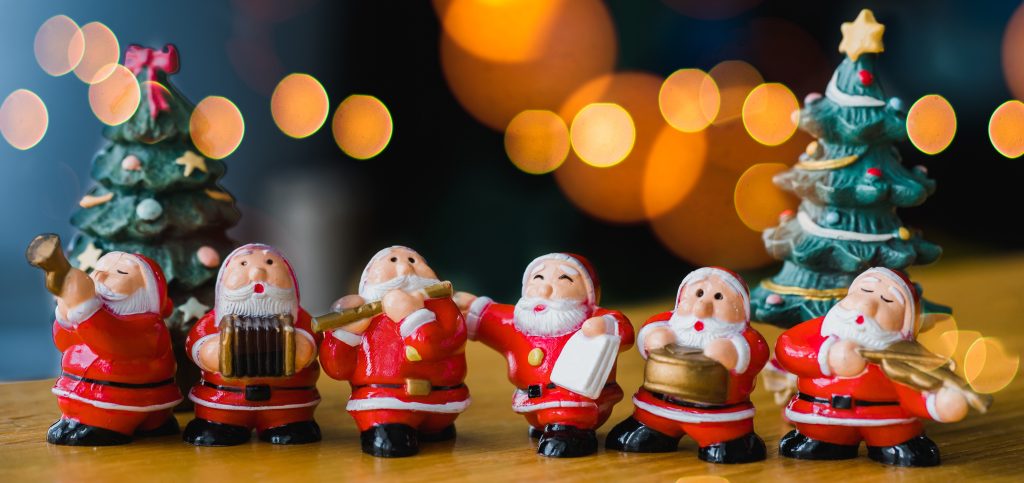 Line of singing and music playing ceramic Santa ornaments one table with Christmas tree decorations and blurred Christmas lights in background