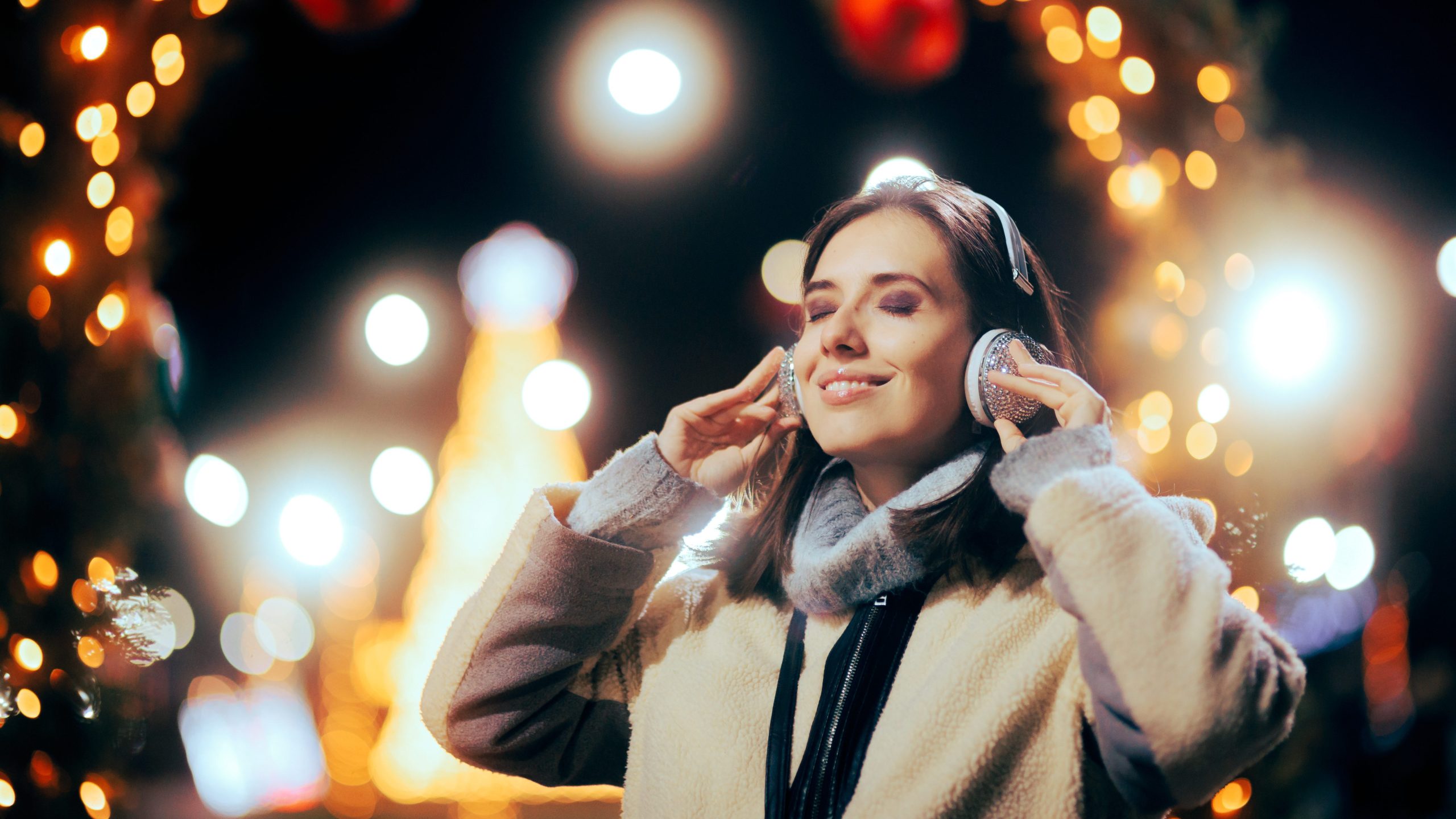 The Golden Age of Holiday Music - Image of a woman standing outside amongst Christmas lights and decorations with headphones on and a calm happy expression on her face, listening to holiday music at a holiday event